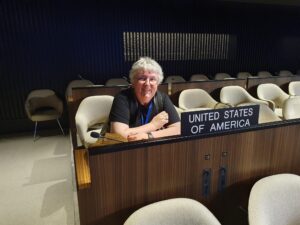 A white woman with glasses is pictured smiling at the camera while sitting at the seat for the United States of America at the UN meeting.