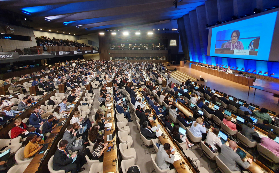A photo of a large meeting space with people sitting in rows looking at a panel of speakers on a stage.